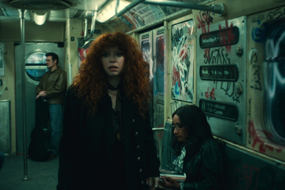 A woman with voluminous curly red hair looks forward in a dimly lit subway car.
