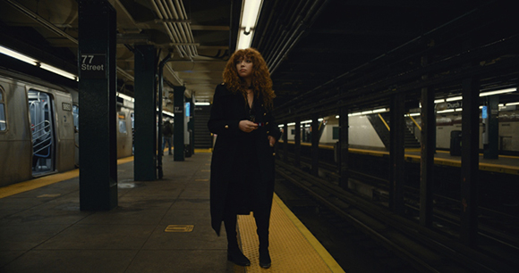 A woman with curly hair wearing a long black coat stands on a dimly lit subway platform.
