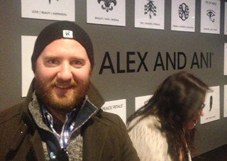 Man with beard and beanie smiles in front of Alex and Ani logo and display of symbols.