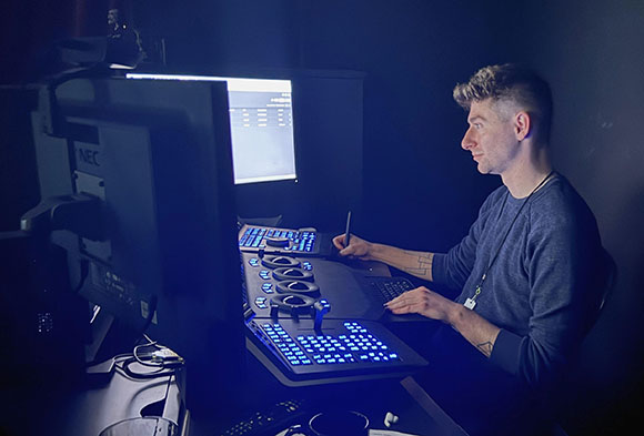 A man sits in a dark room operating a large control console with glowing blue buttons and multiple screens.