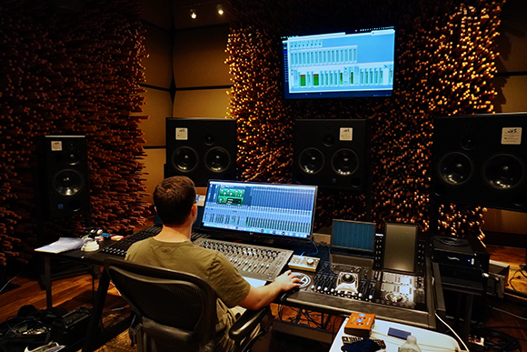 A person sits at a desk with multiple monitors, studio monitors, and a mixing console in a soundproofed room.