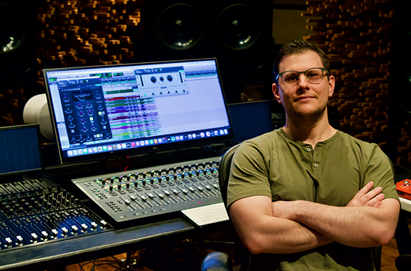 A man with glasses and an olive green shirt sits in a recording studio with arms crossed.