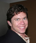 A smiling man with wavy dark hair wearing a dark blazer and a patterned collared shirt.
