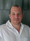 A man with short hair wearing a white and blue striped button-down shirt, smiling against a wooden background.