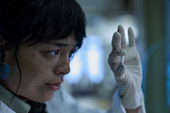 A person wearing a white lab coat and gloves holds a small vial between their thumb and forefinger, looking at it intently. T