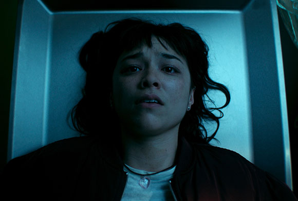 A young woman with dark hair and a distressed expression lies on her back in a blue-lit morgue drawer.