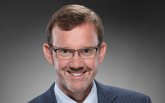 A smiling man with light brown hair and glasses wearing a blue suit jacket and light blue shirt.