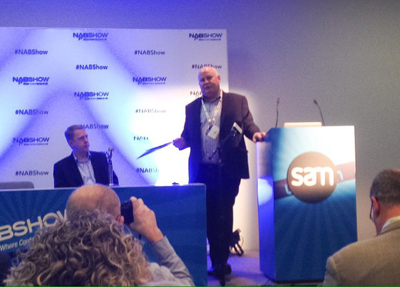 A man in a dark suit stands at a podium labeled SAM, speaking in front of a NABShow branded backdrop.