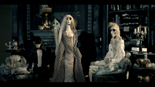 Three pale, stylized figures with long hair and dark clothing stand in a dimly lit, antique-filled room.