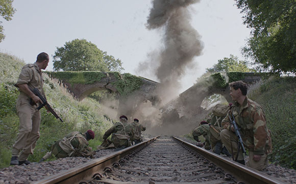 Soldiers in military uniform crouch near railroad tracks as a large plume of smoke rises from a nearby bridge.