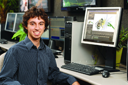 A young man in a striped shirt smiles while sitting at a desk with a computer monitor showing a bee illustration.