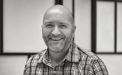 A smiling, bald man with a beard wearing a checkered button-down shirt.