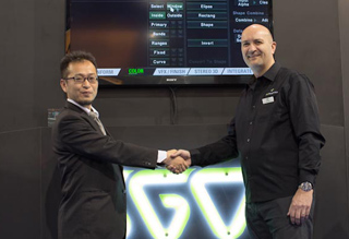 Two men in professional attire shake hands in front of a large computer monitor displaying software menus.