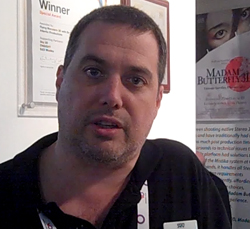 A man with short dark hair and light stubble looking forward in an office.