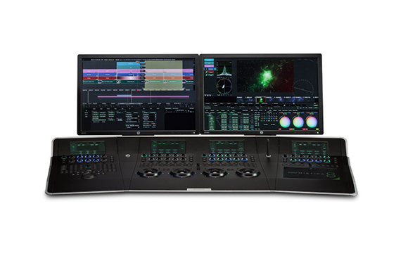 Dual monitors displaying video editing software above a wide black control panel with numerous buttons and dials.