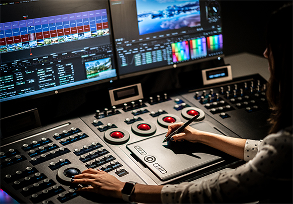 A person's hands operate a video editing control surface with a stylus and knobs in front of dual monitors.