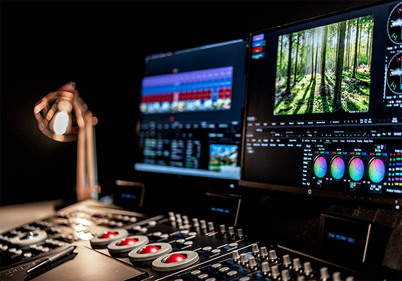Video editing workstation with dual monitors displaying forest footage and color grading tools over a control panel.
