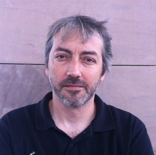 A man with salt-and-pepper hair and a beard wearing a black polo shirt against a textured wall.