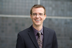 A smiling man with glasses wearing a dark suit, striped shirt, and patterned tie.
