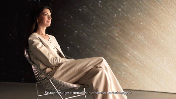 A woman in a cream-colored suit sits in a wire chair against a shimmering, textured wall.