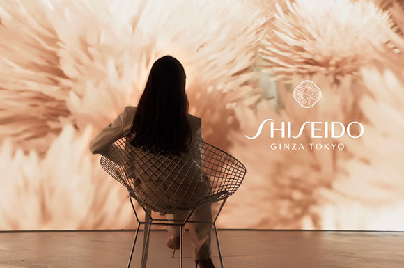A person with long dark hair sits in a wire chair facing a large, soft-focus floral backdrop.