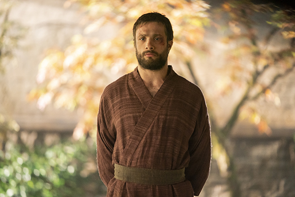 A bearded man in a brown textured robe stands before autumn trees.