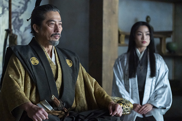 A man in gold and black robes looks sideways while a woman in white sits behind him.