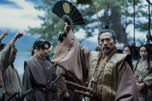 A man in ornate historical Japanese clothing raises a black folding fan during a gathering.