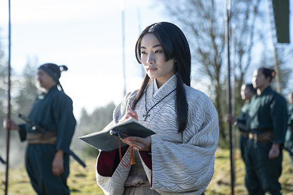 A woman in a patterned light-colored kimono holds an open book while standing in a field with soldiers behind her.