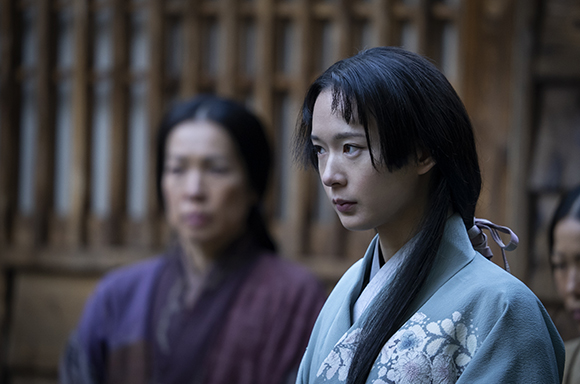 A young woman in light blue traditional attire looks thoughtfully to the side against a blurred wooden background.