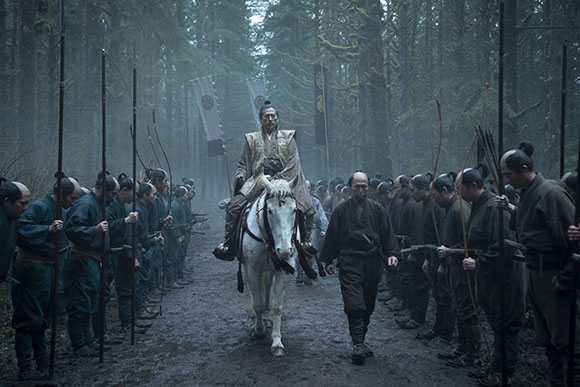A man on a white horse rides through a misty forest flanked by rows of soldiers holding long spears.