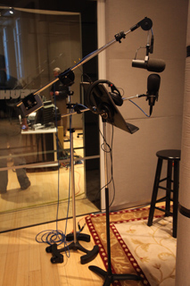 Two microphones on stands with headphones attached, in a recording studio with a glass wall and stool.