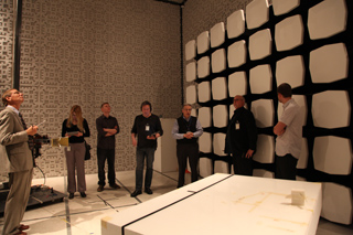 A group of people stand in a room with anechoic panels on the walls, looking at a large white object in the foreground.
