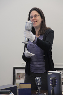 A woman wearing white gloves holds a large silver microphone. Two smaller, similar microphones are on a table.
