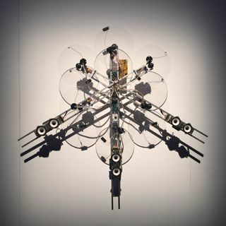 Kinetic sculpture with multiple satellite dishes, speakers, and electronic components arranged radially, casting shadows on a
