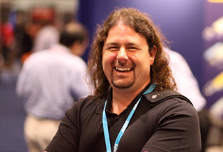 A man with long brown hair and a goatee smiles while wearing a black polo shirt and a blue lanyard.