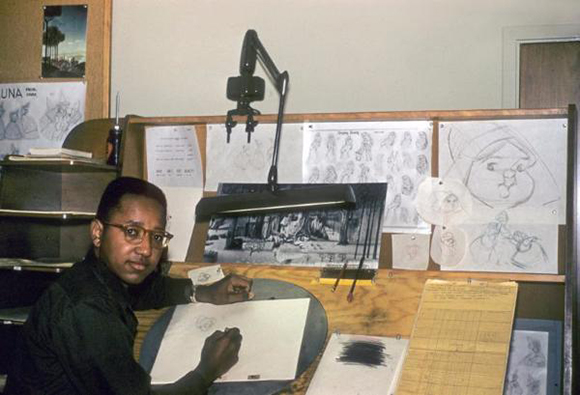A man with glasses sits at a wooden drafting desk covered in sketches and art supplies.