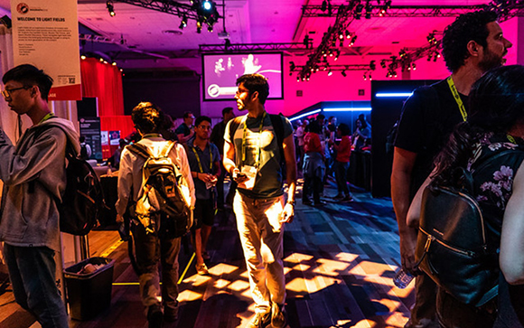 People walking through a convention hall illuminated by pink and blue lights.