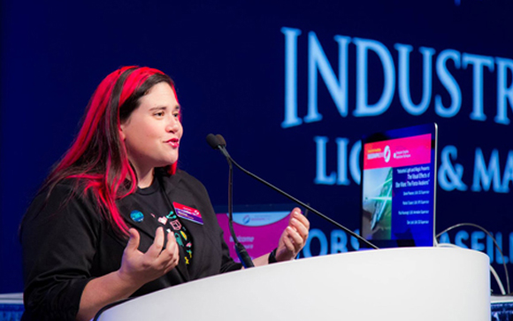 A woman with red-tinted hair gestures with her hands while speaking at a podium during a conference.