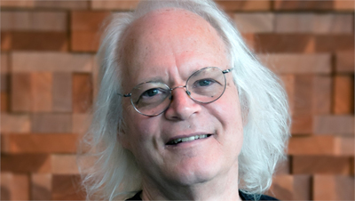 A smiling man with long white hair and round glasses in front of a brick wall.
