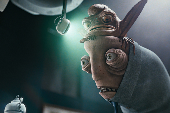 A two-headed, brownish alien creature with large eyes wearing a grey suit looks up toward a hanging light.