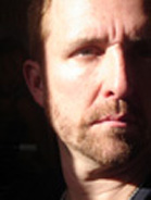 Close-up portrait of a man with light eyes and a short beard looking slightly off-camera.
