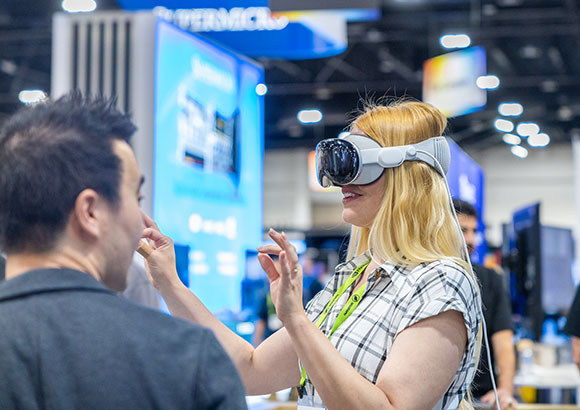 A blonde woman wearing a white VR headset gestures toward a man in a crowded convention hall.