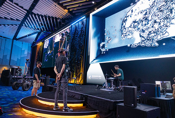 A large digital screen showing blue liquid patterns above a stage with people and technical equipment.