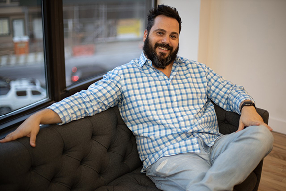 A smiling man with a beard wearing a blue and white checkered shirt sits on a dark tufted sofa.