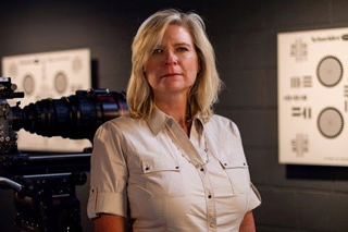 A blonde woman in a light-colored button-down shirt stands in a studio next to a professional film camera.