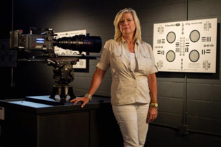 A blonde woman in a tan shirt stands beside a large cinema camera mounted on a tripod in a dark studio.