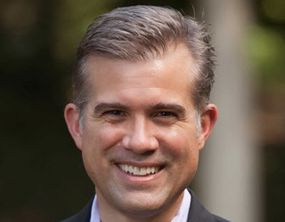 A smiling man with short, salt-and-pepper hair wearing a dark blazer and light purple shirt.