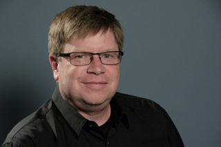A man with short brown hair and glasses wearing a black button-down shirt against a gray background.