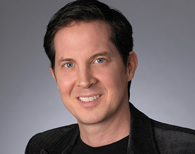 A man with short dark hair and blue eyes smiling in a black blazer and black shirt.